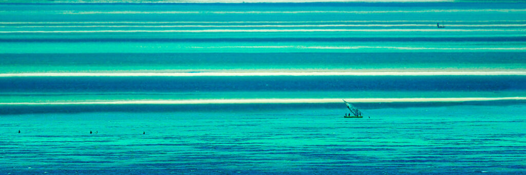 Photographie « Mozambique », série Horizons de Sabine Chabot. Dans les bandes de lumière et de turquoise du lagon, une embarcation et quelques silhouettes ponctuent l’immensité